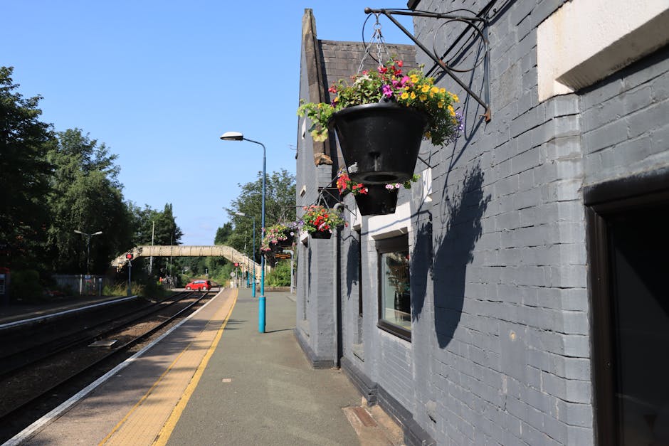 A daytime view of a train station platform adjacent to a grey brick building featuring black hanging flower baskets filled with colorful pink, yellow, purple, and white blooms, set against a clear blu