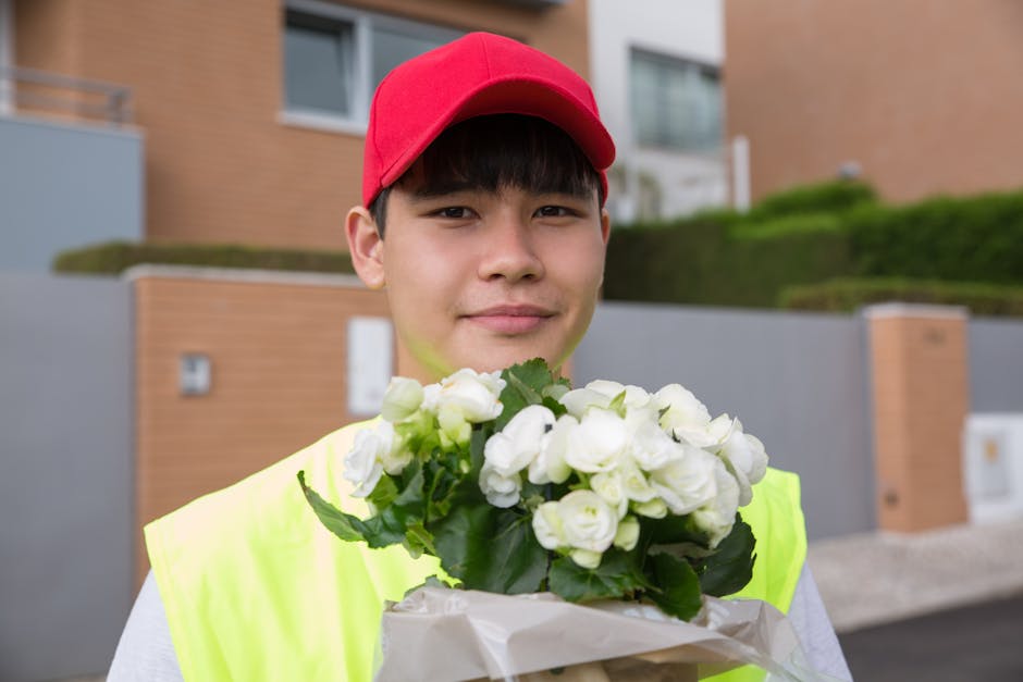 A young delivery person wearing a red cap and high-visibility yellow vest holds a bouquet of fresh white roses and ranunculus with green leaves, wrapped in white tissue paper. The person stands outdoo